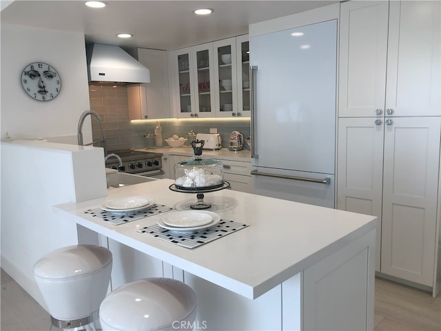 kitchen with built in fridge, wall chimney range hood, white cabinets, and a kitchen bar