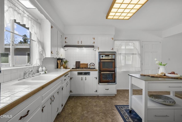 kitchen featuring tile counters, multiple ovens, decorative backsplash, sink, and white cabinets