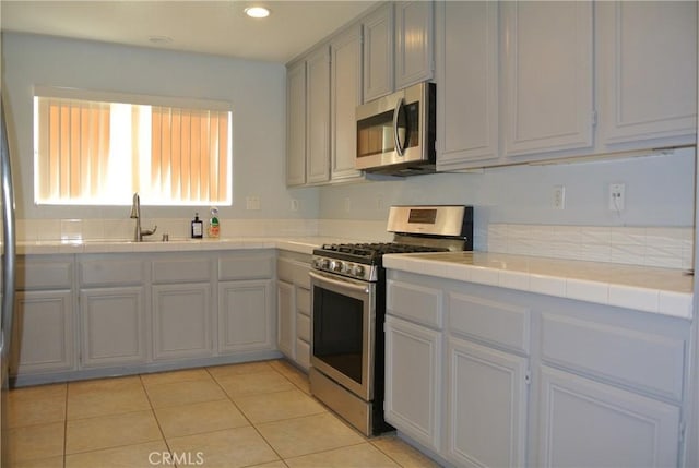 kitchen with light tile patterned flooring, sink, tile counters, gray cabinets, and stainless steel appliances