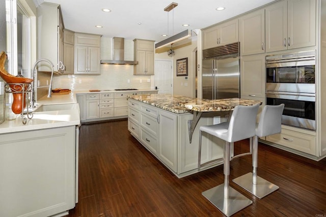 kitchen with dark wood-style floors, a kitchen island, a sink, stainless steel appliances, and wall chimney range hood