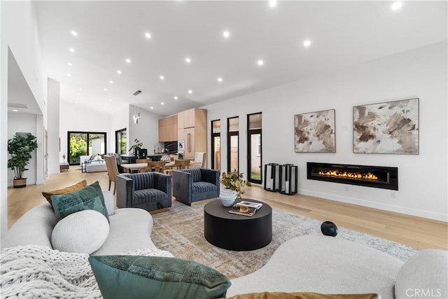 living room featuring light wood finished floors, baseboards, a glass covered fireplace, recessed lighting, and high vaulted ceiling
