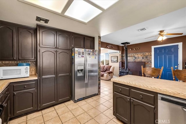 kitchen with appliances with stainless steel finishes, light tile patterned floors, dark brown cabinets, a wood stove, and ceiling fan