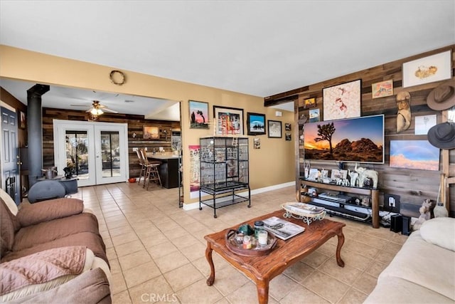 living room with french doors, wooden walls, a wood stove, ceiling fan, and light tile patterned floors