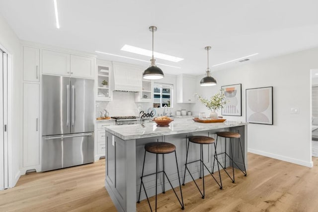 kitchen featuring a skylight, hanging light fixtures, stainless steel fridge, an island with sink, and white cabinets