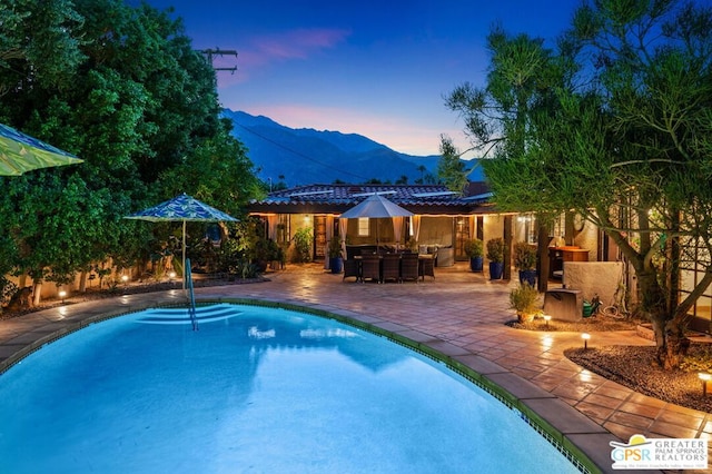 pool at dusk featuring an outdoor fireplace, a mountain view, and a patio area