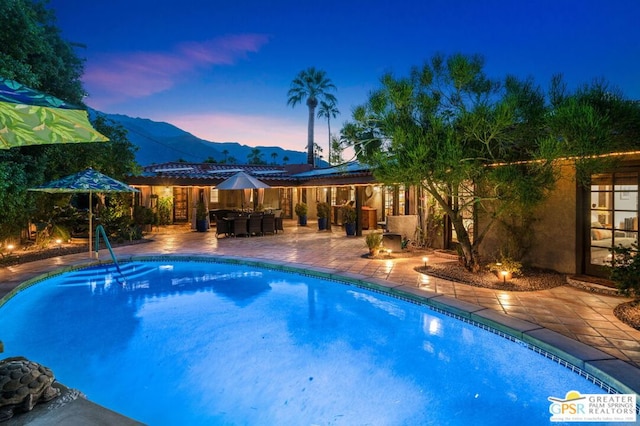 pool at dusk with a mountain view, a patio area, and an outdoor bar