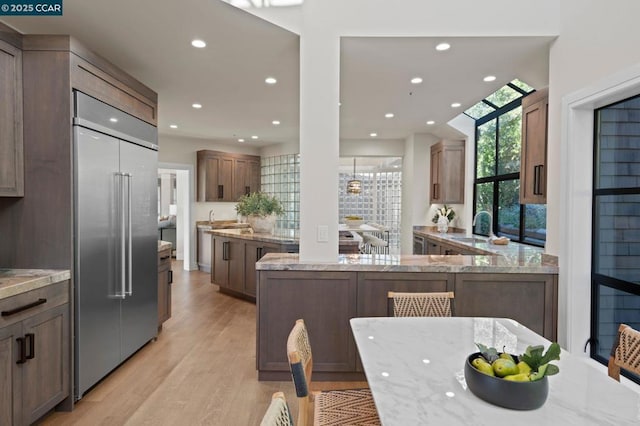 kitchen with light stone countertops, stainless steel built in refrigerator, sink, kitchen peninsula, and light hardwood / wood-style flooring