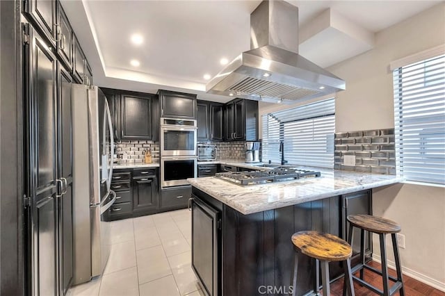 kitchen featuring light stone countertops, island exhaust hood, stainless steel appliances, a kitchen breakfast bar, and kitchen peninsula