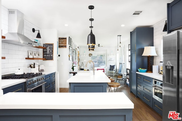 kitchen with appliances with stainless steel finishes, dark hardwood / wood-style floors, wall chimney range hood, an island with sink, and decorative backsplash