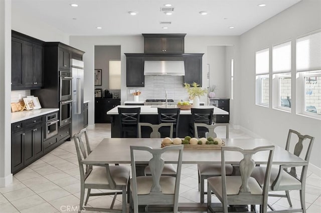 kitchen featuring backsplash, a kitchen breakfast bar, a kitchen island with sink, stainless steel double oven, and wall chimney exhaust hood