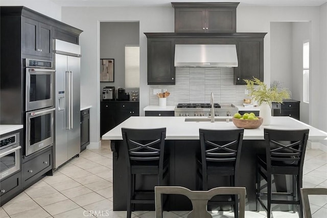 kitchen featuring a breakfast bar area, light tile patterned floors, appliances with stainless steel finishes, an island with sink, and exhaust hood