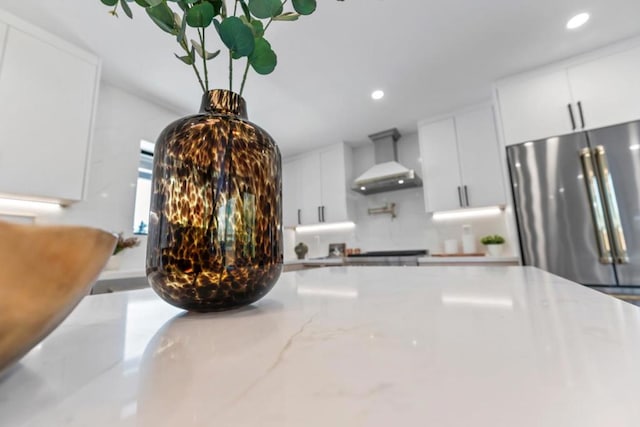 interior details with high end refrigerator, tasteful backsplash, white cabinetry, light stone counters, and wall chimney exhaust hood