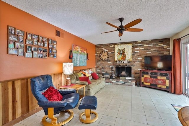 tiled living room featuring ceiling fan, wooden walls, a brick fireplace, and a textured ceiling