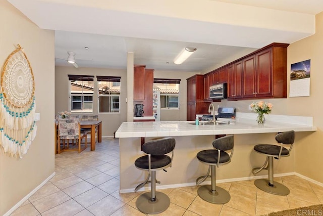 kitchen with reddish brown cabinets, stainless steel range with gas cooktop, tile countertops, light tile patterned flooring, and a peninsula