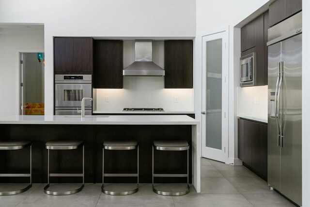 kitchen featuring a kitchen breakfast bar, dark brown cabinets, wall chimney exhaust hood, sink, and built in appliances