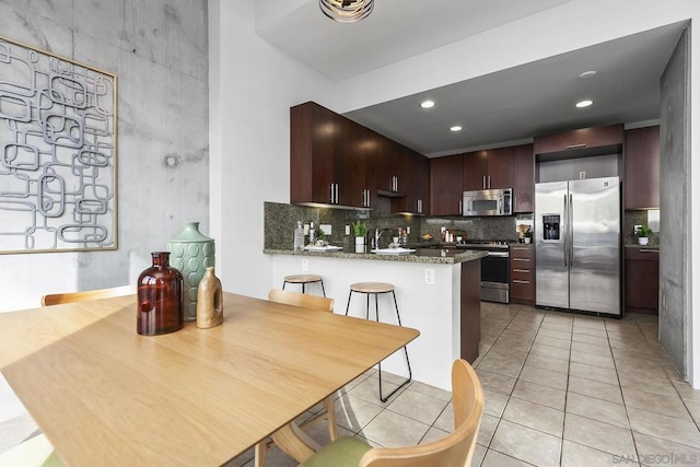 kitchen featuring appliances with stainless steel finishes, stone countertops, kitchen peninsula, light tile patterned floors, and dark brown cabinets