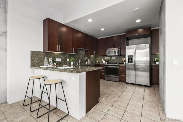 kitchen featuring stainless steel appliances, decorative backsplash, kitchen peninsula, light tile patterned floors, and dark brown cabinets