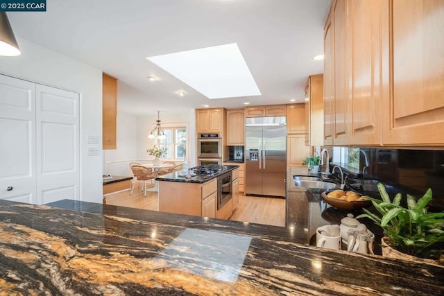 kitchen featuring appliances with stainless steel finishes, a skylight, dark stone counters, light brown cabinetry, and sink