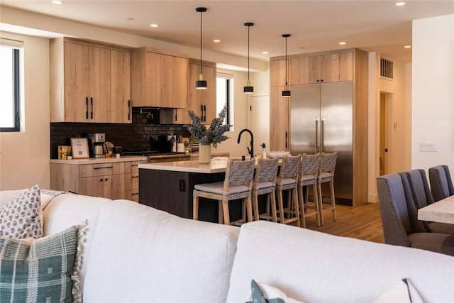 kitchen with pendant lighting, stainless steel built in refrigerator, an island with sink, backsplash, and light brown cabinets