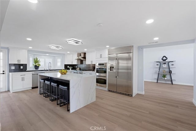 kitchen with wall chimney range hood, white cabinetry, a kitchen island, stainless steel appliances, and a kitchen bar