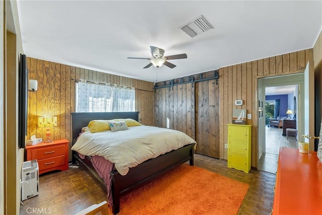 bedroom featuring dark parquet floors, wooden walls, ceiling fan, and a barn door