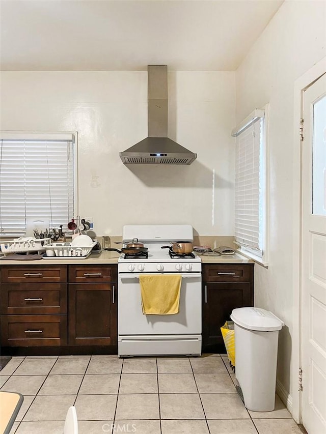 kitchen featuring wall chimney range hood, dark brown cabinetry, white gas stove, and light tile patterned floors