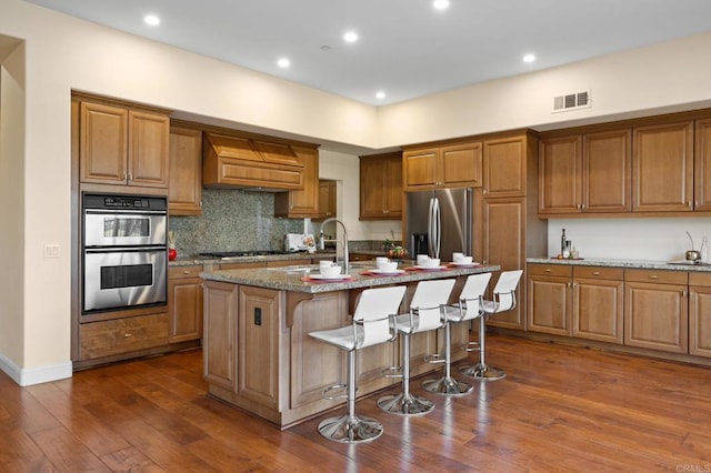 kitchen with a kitchen breakfast bar, custom range hood, stainless steel appliances, light stone countertops, and a kitchen island with sink