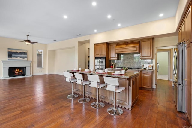 kitchen featuring appliances with stainless steel finishes, a kitchen island with sink, tasteful backsplash, light stone countertops, and custom range hood