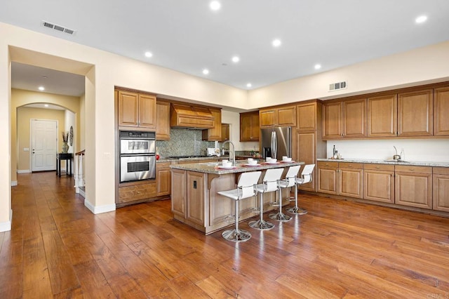 kitchen featuring a breakfast bar, appliances with stainless steel finishes, wood-type flooring, light stone countertops, and an island with sink