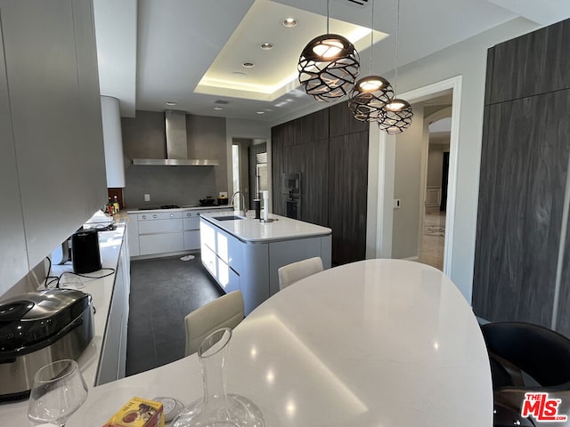 kitchen featuring hanging light fixtures, wall chimney exhaust hood, sink, white cabinets, and a center island with sink