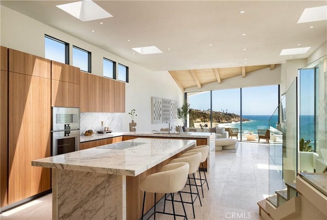 kitchen featuring a water view, vaulted ceiling with beams, backsplash, light stone counters, and a breakfast bar area