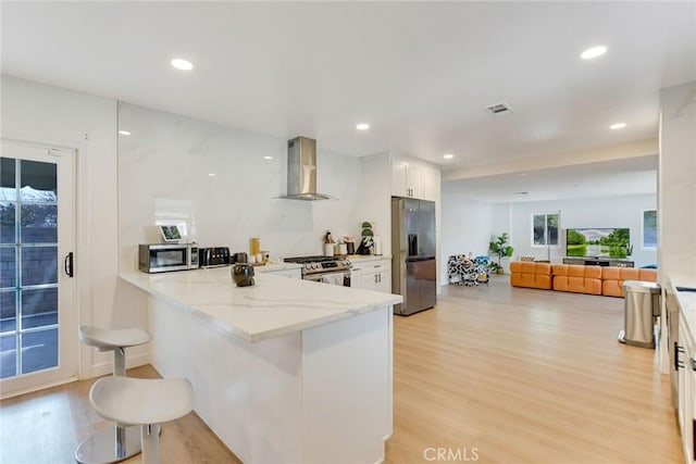 kitchen featuring light stone countertops, white cabinets, kitchen peninsula, stainless steel appliances, and wall chimney exhaust hood