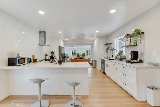 kitchen featuring wall chimney range hood, stainless steel appliances, white cabinets, and kitchen peninsula