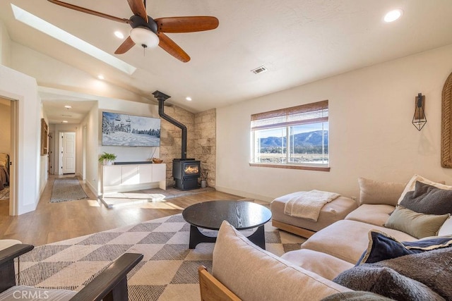 living room with light wood-type flooring, a wood stove, lofted ceiling with skylight, and ceiling fan