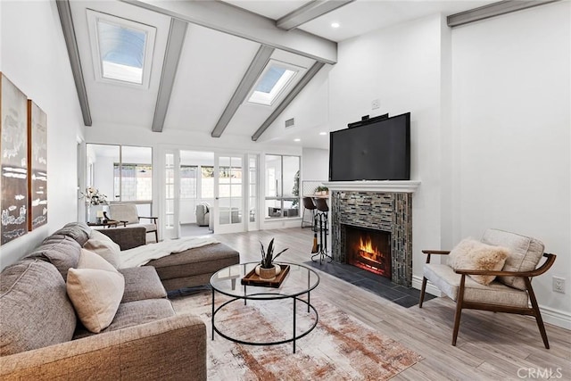 living room featuring high vaulted ceiling, a skylight, a tiled fireplace, beam ceiling, and light hardwood / wood-style flooring