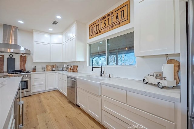 kitchen featuring light stone countertops, white cabinets, wall chimney range hood, stainless steel appliances, and light hardwood / wood-style floors