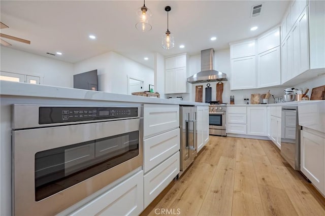 kitchen featuring hanging light fixtures, appliances with stainless steel finishes, wall chimney exhaust hood, light wood-type flooring, and white cabinetry