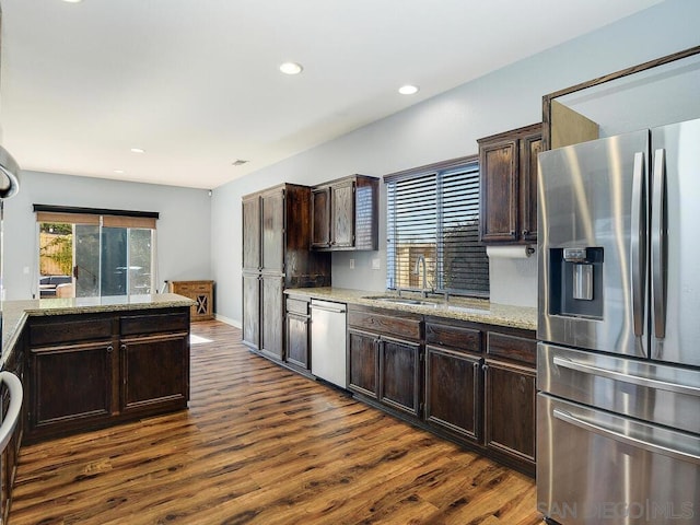 kitchen with appliances with stainless steel finishes, sink, dark brown cabinets, dark hardwood / wood-style flooring, and light stone counters