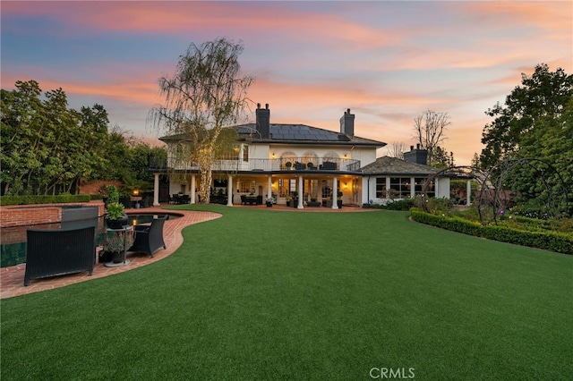 back of house at dusk featuring a lawn, a patio, roof mounted solar panels, a balcony, and a chimney