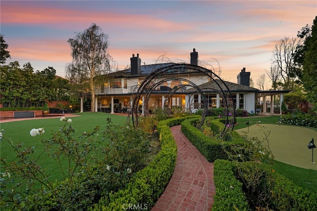 back of house at dusk with a lawn, a chimney, and a balcony