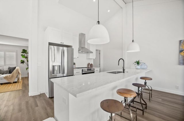 kitchen with wall chimney range hood, sink, stainless steel appliances, white cabinets, and decorative light fixtures