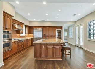 kitchen with an island with sink, sink, exhaust hood, stainless steel appliances, and light stone countertops