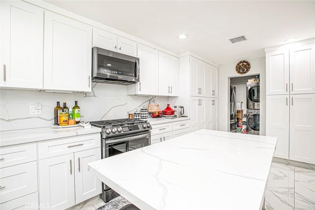 kitchen with appliances with stainless steel finishes, white cabinets, light stone countertops, stacked washer / dryer, and decorative backsplash