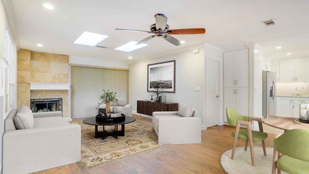 living room with ceiling fan, crown molding, a tiled fireplace, and light wood-type flooring