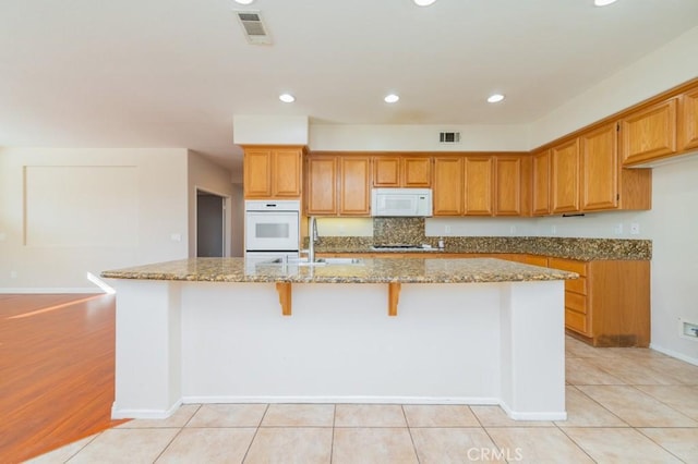 kitchen featuring sink, white appliances, a breakfast bar area, a kitchen island with sink, and light stone countertops