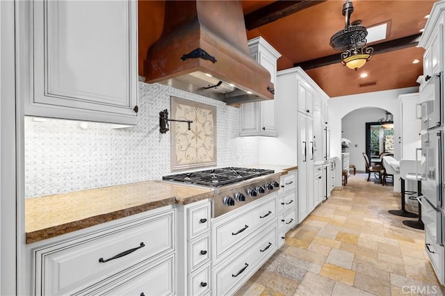 kitchen with custom exhaust hood, white cabinetry, stainless steel gas stovetop, beam ceiling, and decorative backsplash