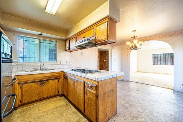 kitchen with stainless steel gas stovetop, tile counters, hanging light fixtures, sink, and kitchen peninsula