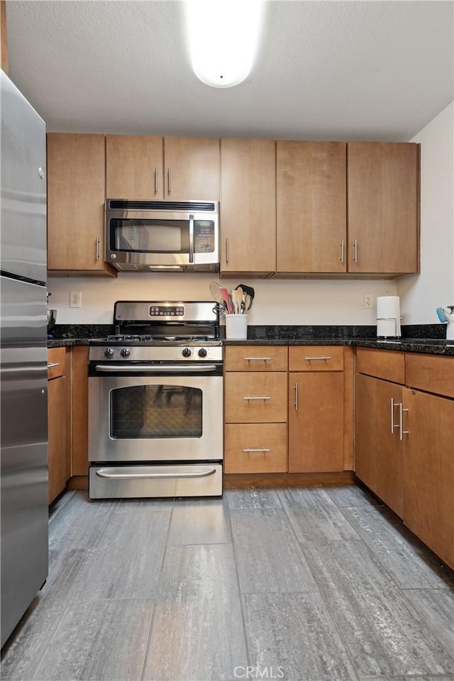 kitchen with light wood-style flooring, appliances with stainless steel finishes, brown cabinets, and dark stone counters