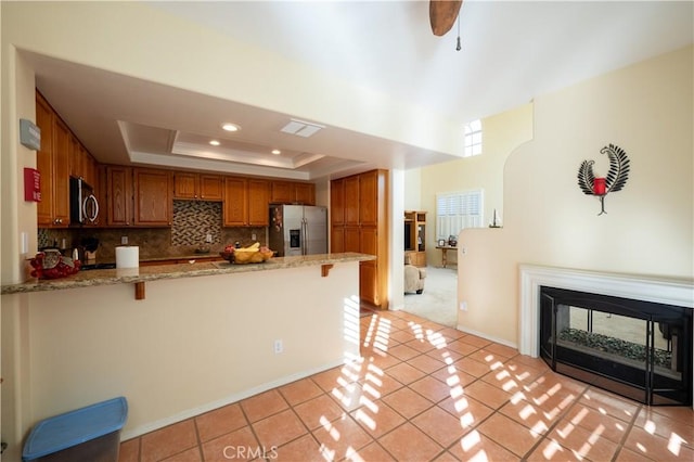 kitchen featuring a multi sided fireplace, appliances with stainless steel finishes, a tray ceiling, light stone counters, and kitchen peninsula