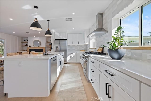 kitchen featuring appliances with stainless steel finishes, pendant lighting, wall chimney range hood, a kitchen island with sink, and white cabinets